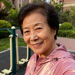 一位五十五岁左右的中国女性，穿着粉色运动外套，在社区健身器材旁微笑看向镜头
